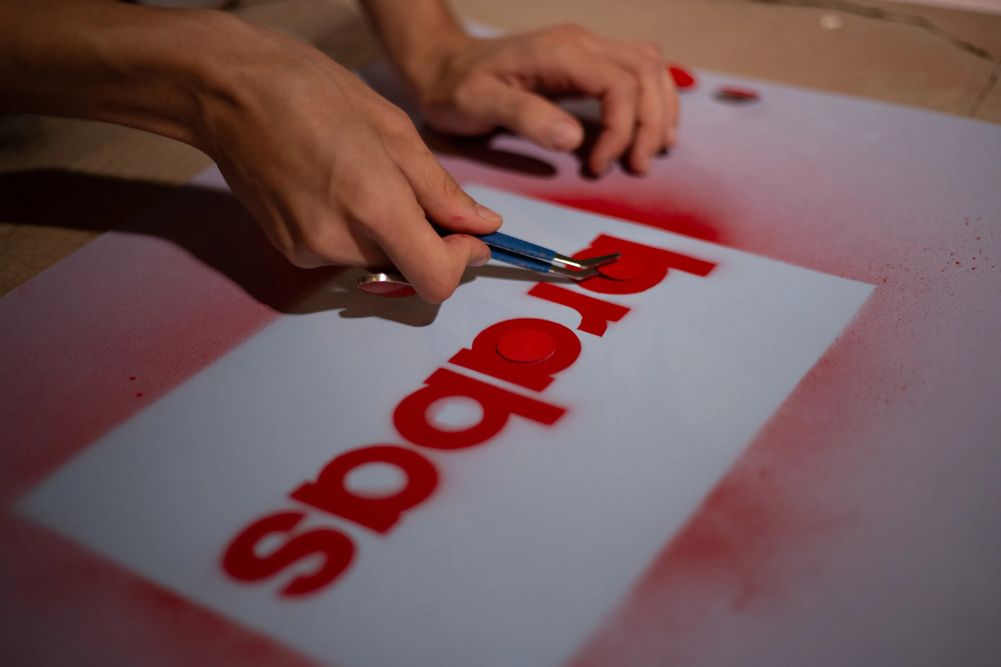 a hand taking the stencil out of the paper with tweezers, on the paper is the adidas logo made with red spray paint